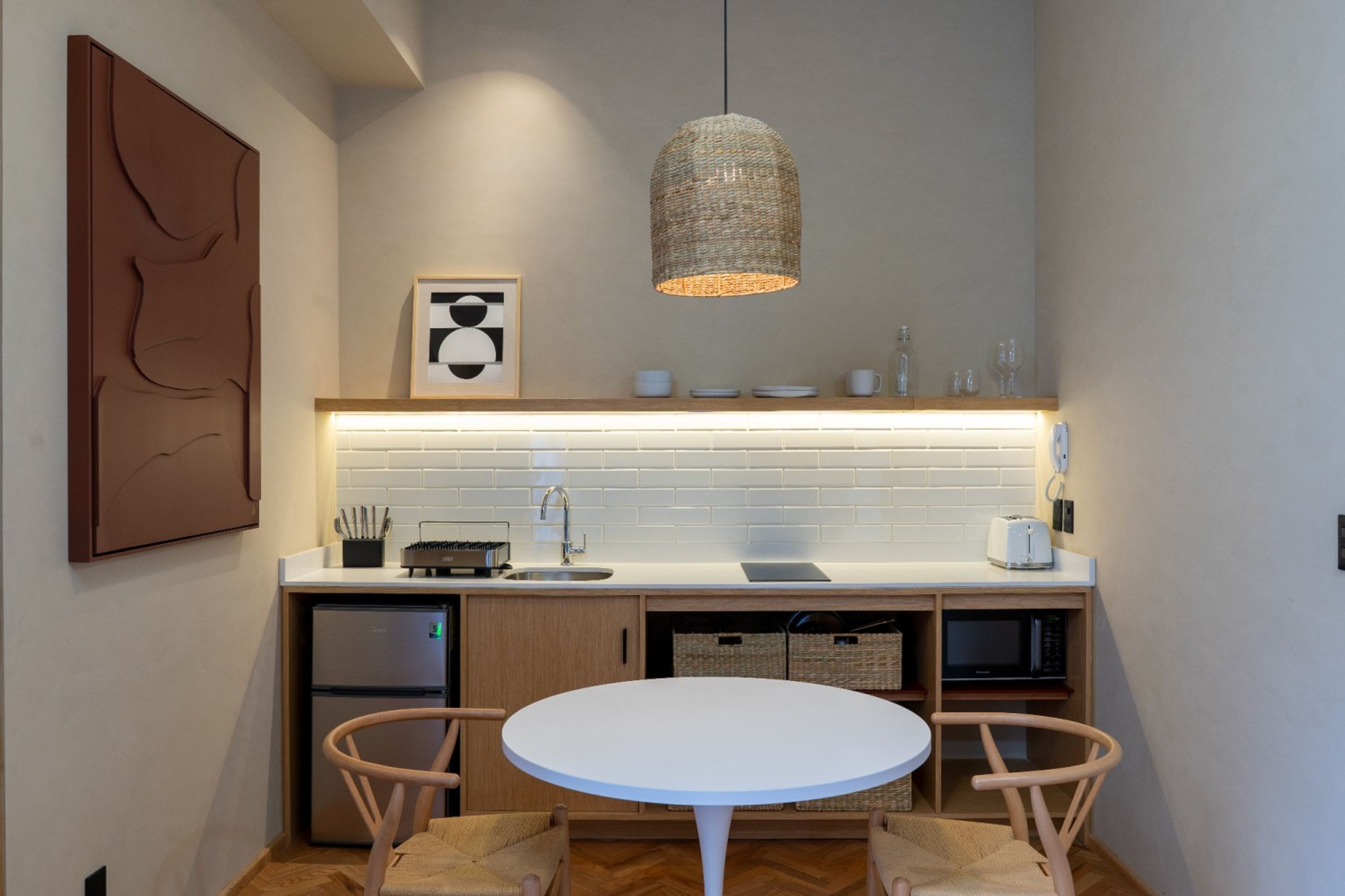 A compact kitchen with a white round table, wooden chairs, a hanging woven light fixture, a microwave, and a refrigerator.