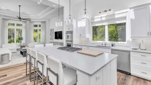 Una cocina moderna con armarios blancos, una isla central, iluminación colgante y ventanas amplias.