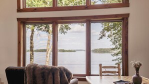 A room with a large window overlooking a lake, a wooden table, a chair, and a vase with flowers. - Moose Lodge/ Firepit/ Swim/ Pontoon/ Kayaks (Pequot Lakes)