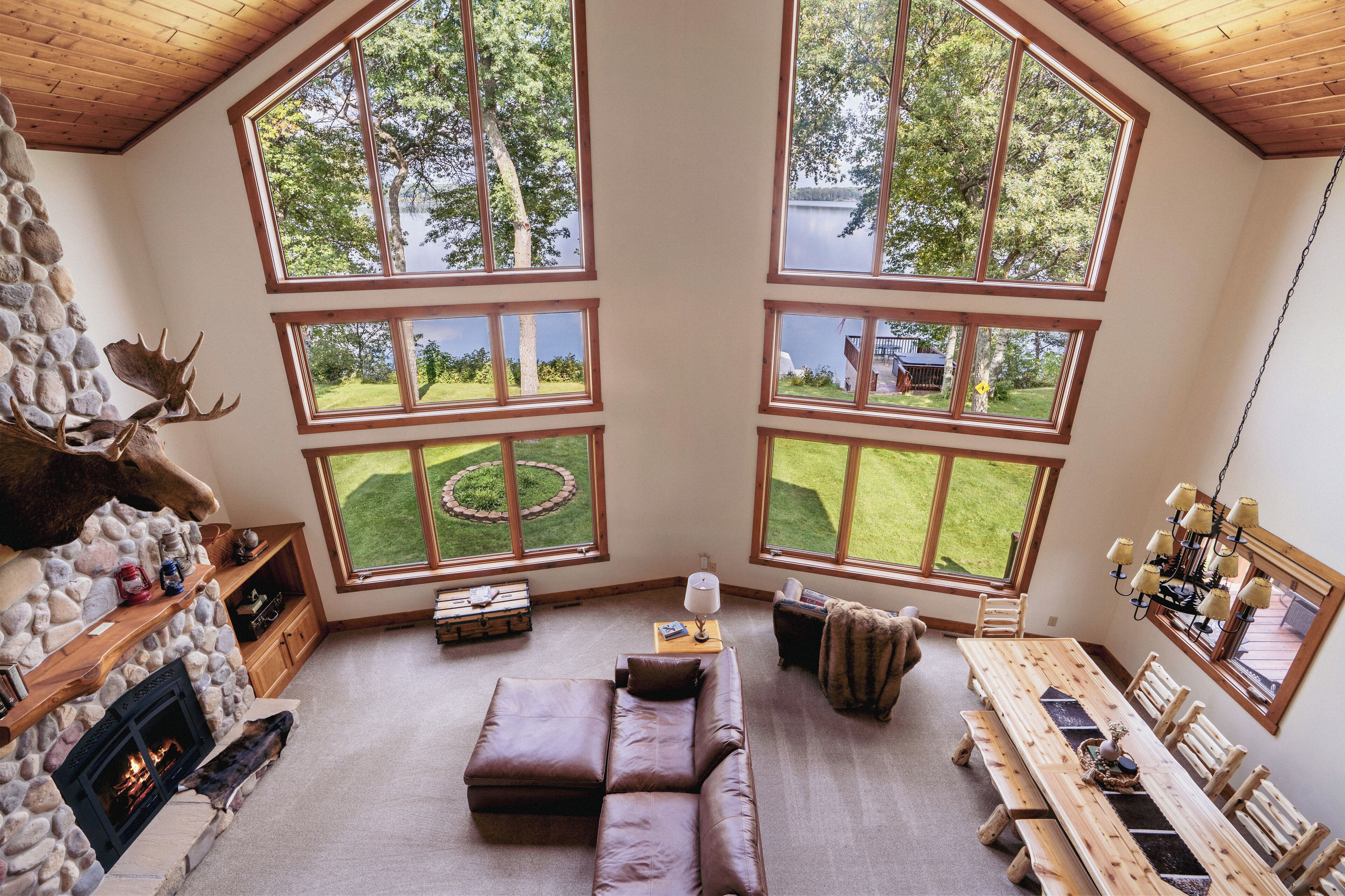 A cozy room with a stone fireplace, a leather sofa, a wooden dining table, and large windows overlooking a lake and greenery.