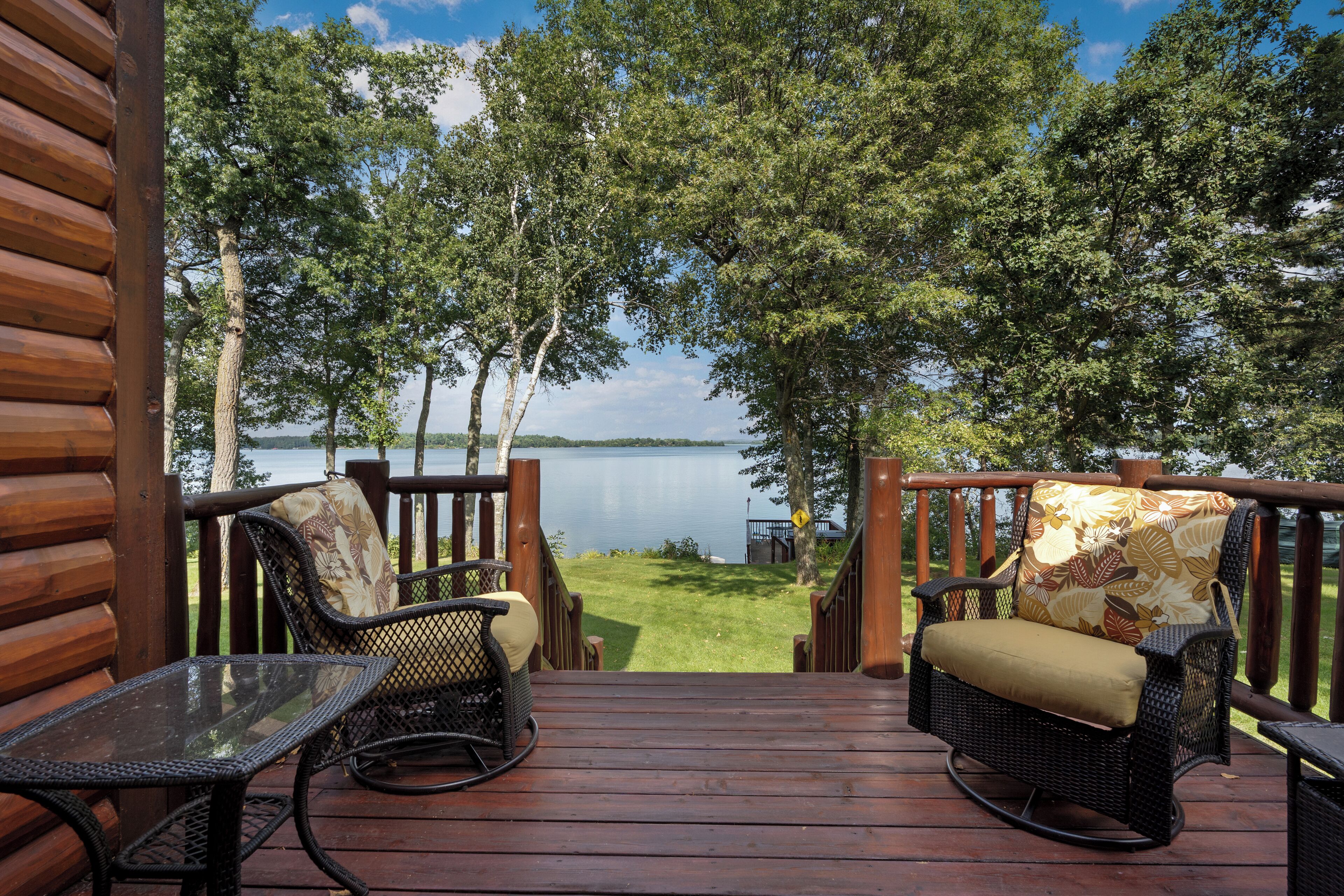 Una terraza de madera con sillas de mimbre y una mesa de vidrio, con vistas a un lago rodeado de árboles.