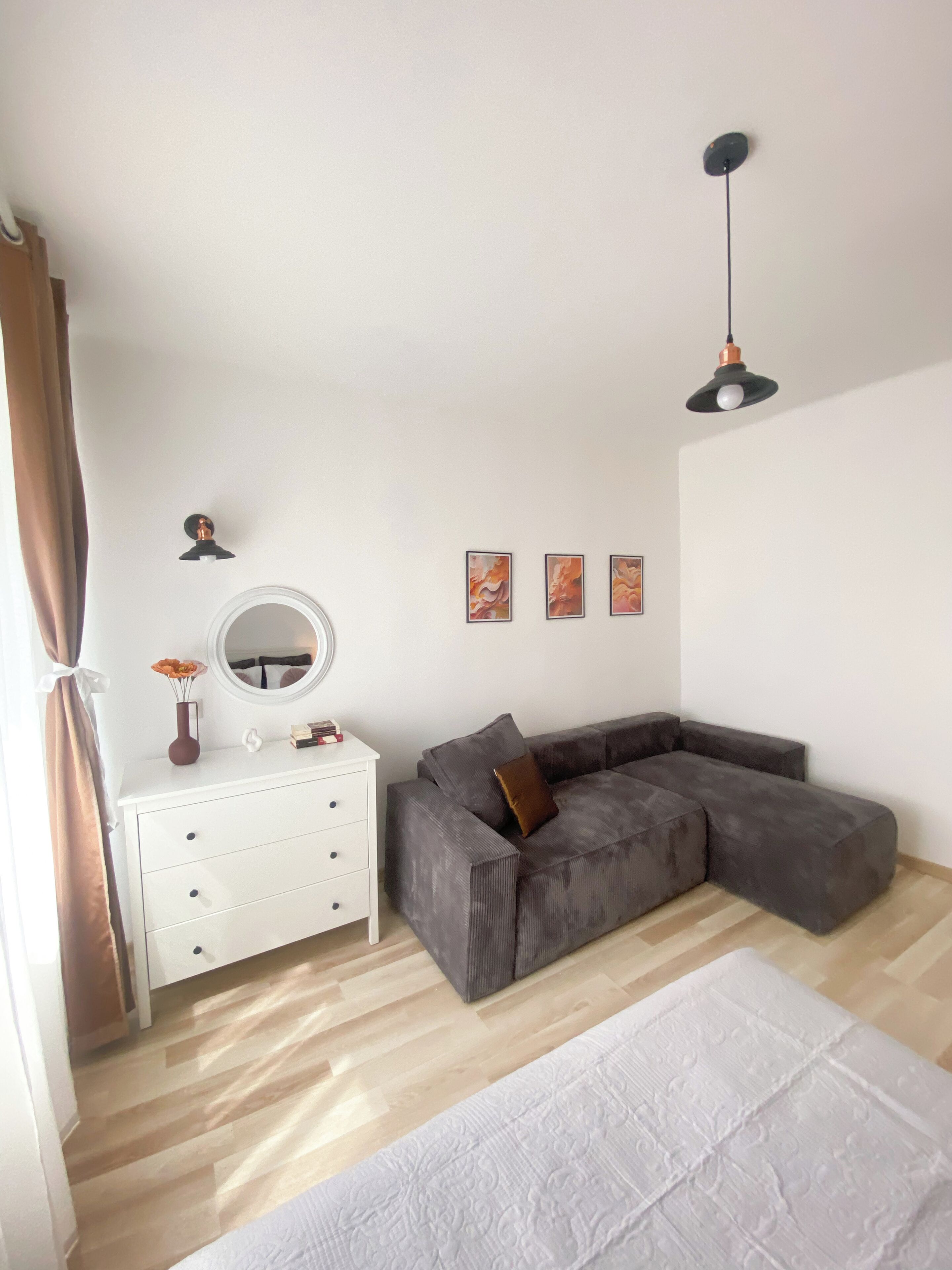 Ein modernes Wohnzimmer mit einem grauen Ecksofa, einer weißen Kommode, einem runden Spiegel und einer hängenden Pendelleuchte.