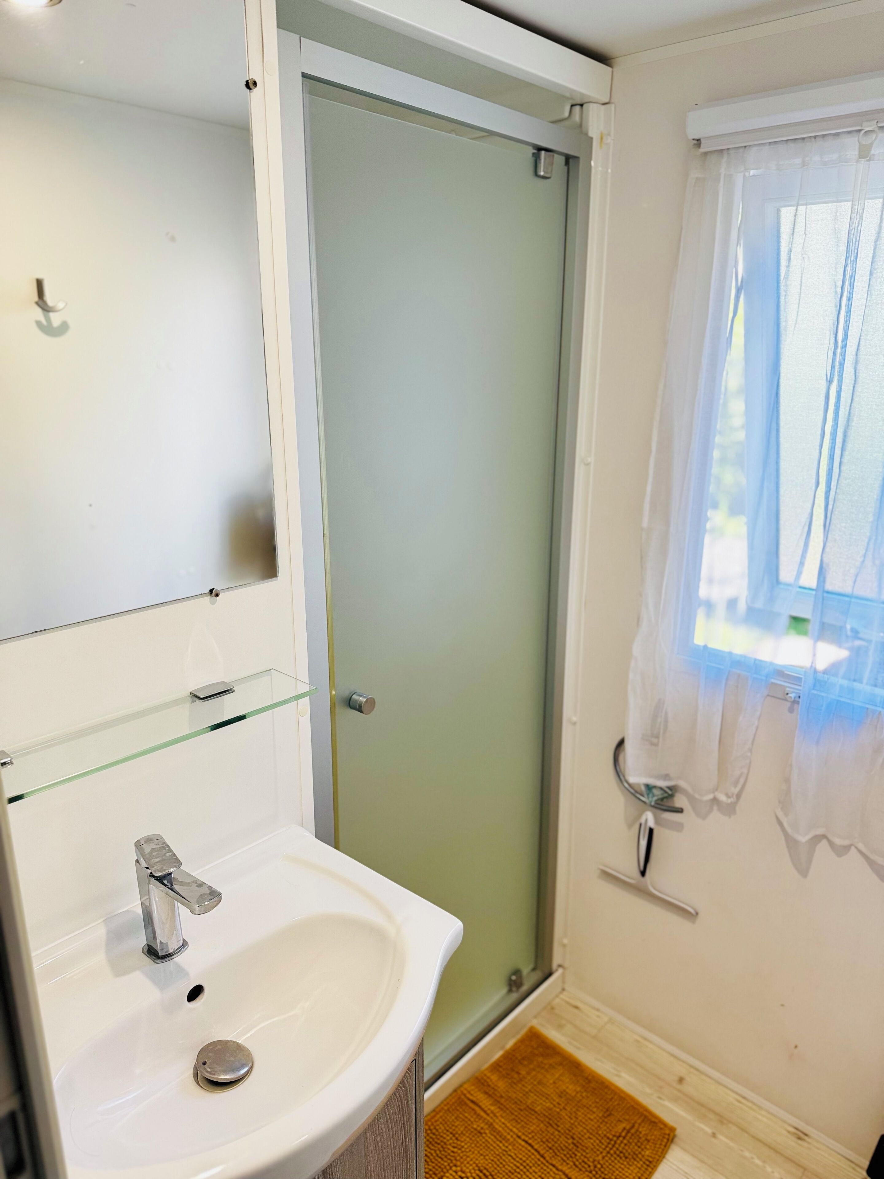 Une salle de bain avec une douche à parois de verre, un lavabo blanc et une fenêtre ornée d’un rideau léger.