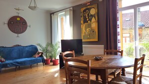 A living room with a blue sofa, wooden dining table, chairs, a TV, potted plants, and a large abstract painting. - Large suite with access to living room, terrace, garden - Thiers center, train station (Thiers)