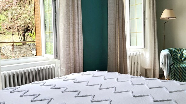 A bedroom with a large bed, a window with curtains, a radiator, and a lamp. - Large suite with access to living room, terrace, garden - Thiers center, train station (Thiers)
