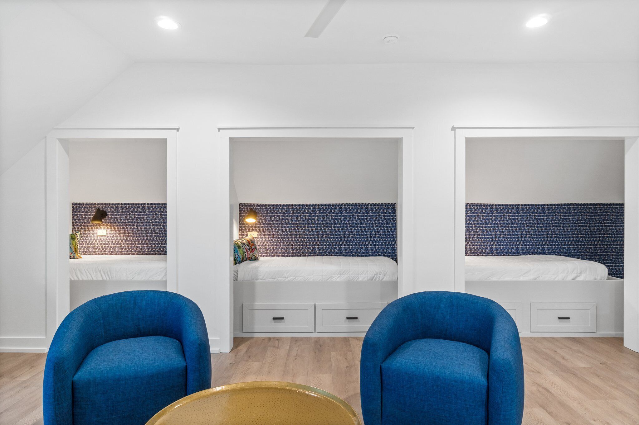 Ein modernes Zimmer mit drei Betten, jedes mit weißem Kopfteil und einer Wand mit Muster. Im Vordergrund stehen zwei blaue Stühle und ein kleiner Tisch.