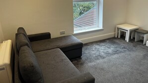 Um quarto com um sofá modular cinza, uma pequena mesa branca e uma janela com vista para um telhado e vegetação.