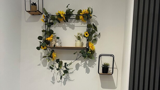 Canto de um quarto com parede branca, um espelho com moldura preta, uma pequena prateleira com uma planta em vaso e um arranjo decorativo de girassóis e folhagem.