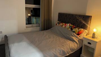 Una cama bien hecha con edredón gris, un cojín colorido y un cabecero tapizado negro. Se ve una mesita de noche con una lámpara y un cajón pequeño. Se divisa una ventana con cortinas que muestra un edificio exterior.
