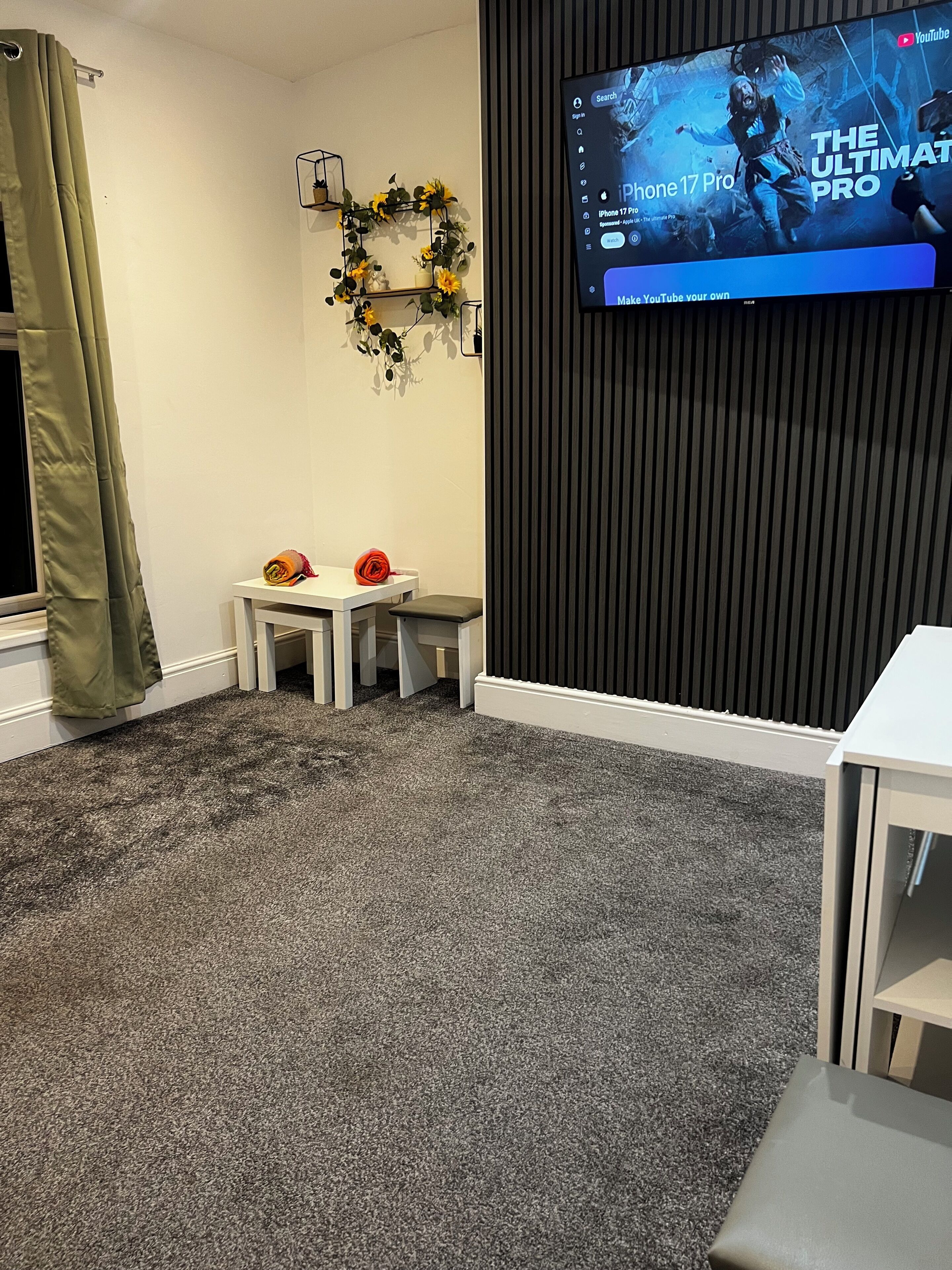 A room with a grey carpet, a white table, two small stools, a TV mounted on a dark wall, and a green curtain.