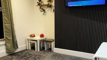 A room with a grey carpet, a white table, two small stools, a TV mounted on a dark wall, and a green curtain.