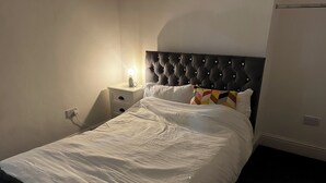 Cama bem arrumada com cabeceira estofada, mesa de cabeceira com abajur e tomada na parede.