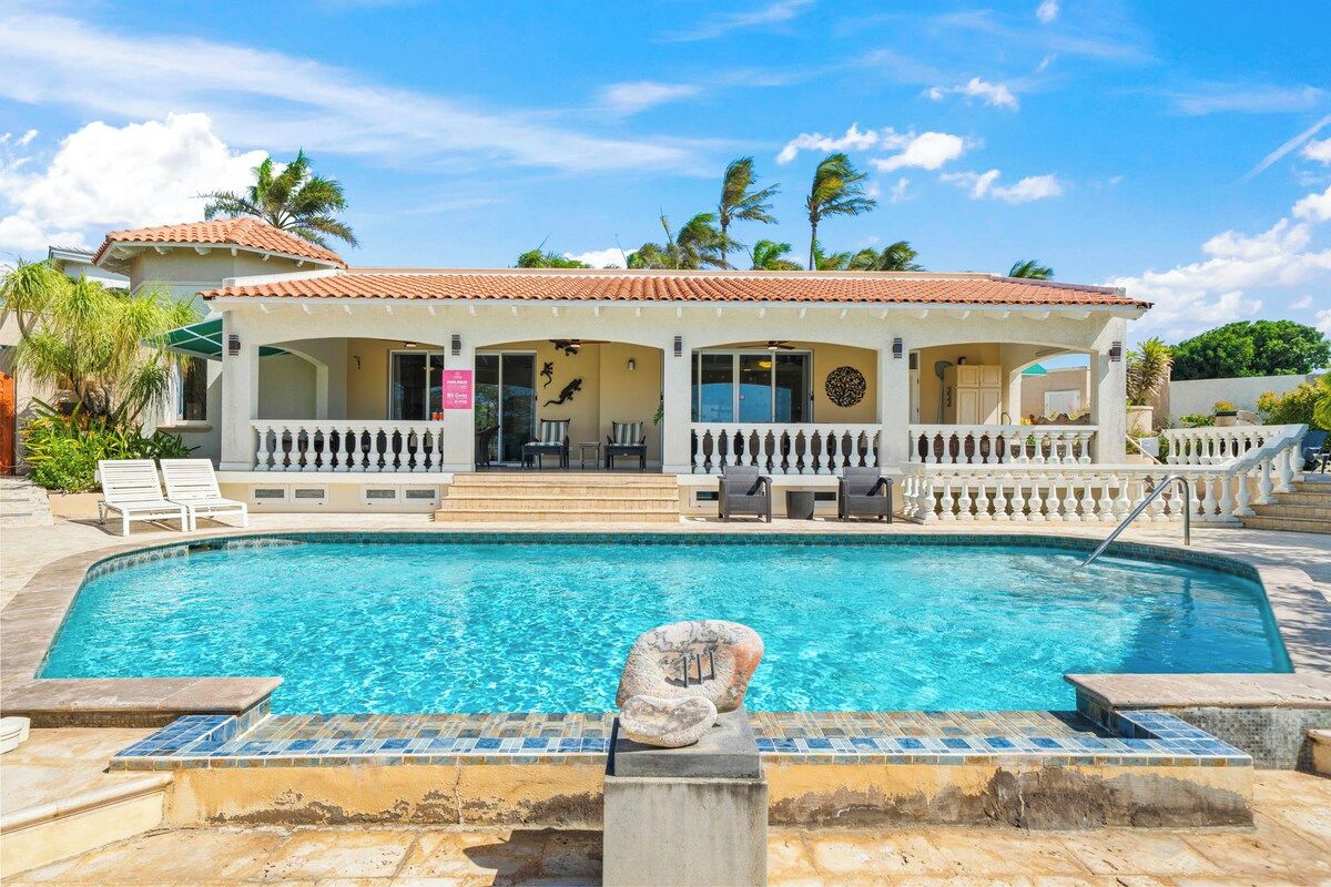 A poolside area with a clear pool, a covered seating area with chairs, and a building with a tiled roof.