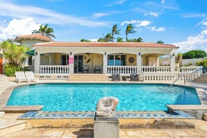 A poolside area with a clear pool, a covered seating area with chairs, and a building with a tiled roof.