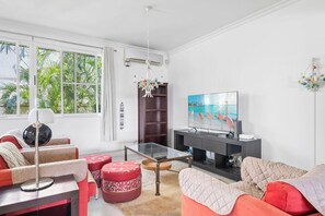 A modern living room with a television, red and beige sofas, a glass coffee table, and a view of palm trees through large windows.