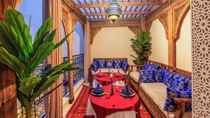 Een traditionele Marokkaanse eethoek met een houten tafel gedekt voor een maaltijd, een hanglamp en een bank met gekleurde kussens.