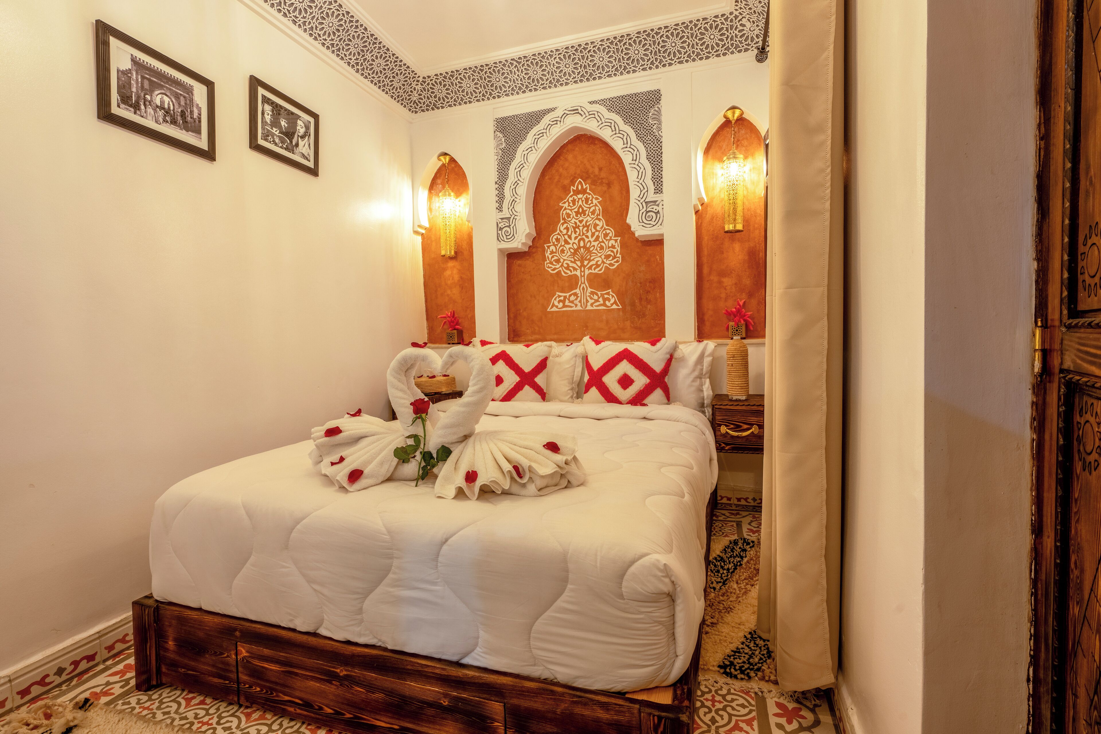 Una cama bien hecha con sábanas blancas y almohadillas decorativas rojas, cabecera de madera con diseño de árbol tallado, y dos luces fijadas a la pared.