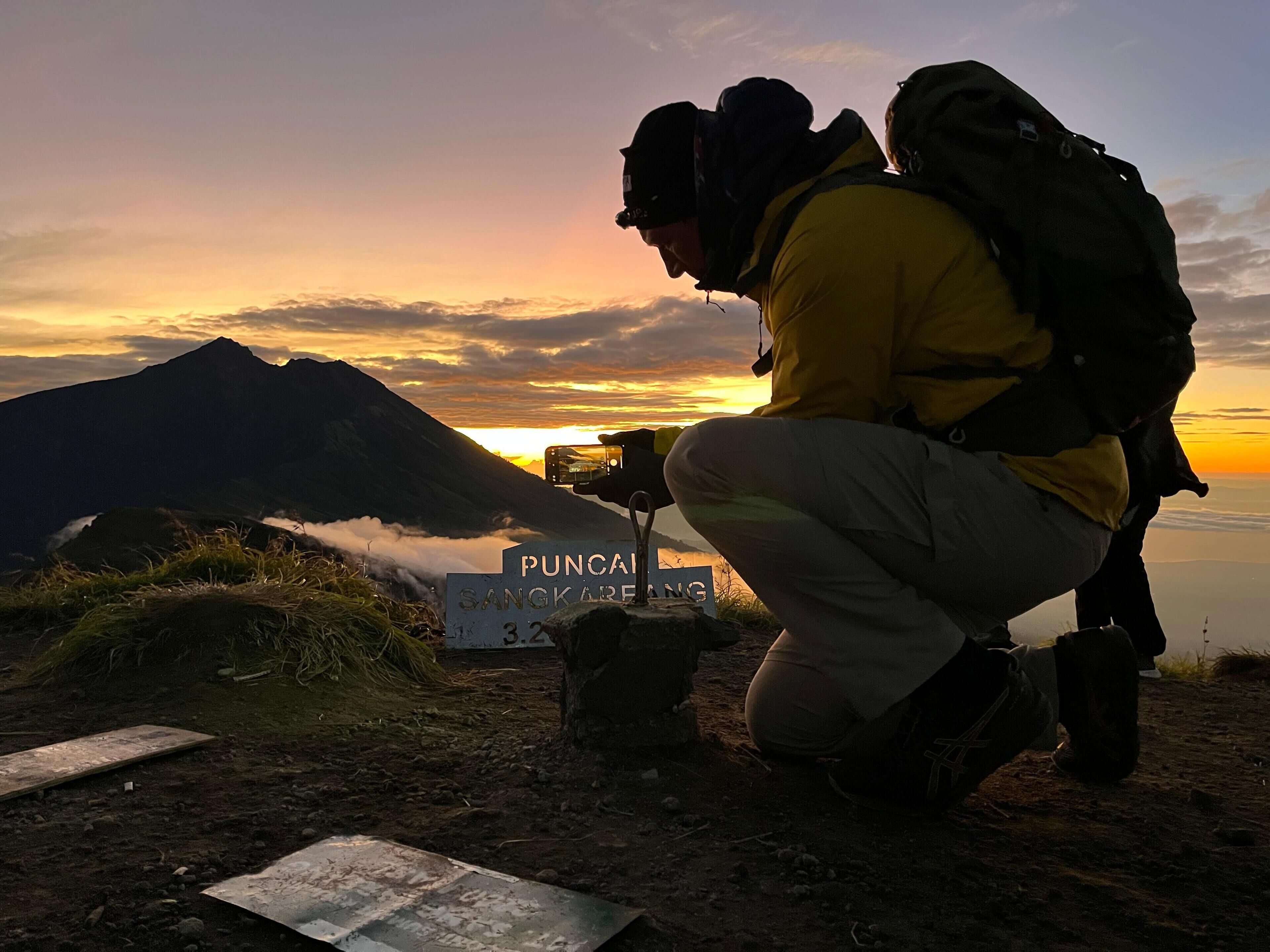 Une personne accroupie sur un sentier de montagne, avec un panneau indiquant « PUNCARANG SANGKARANG », et une montagne en arrière-plan.
