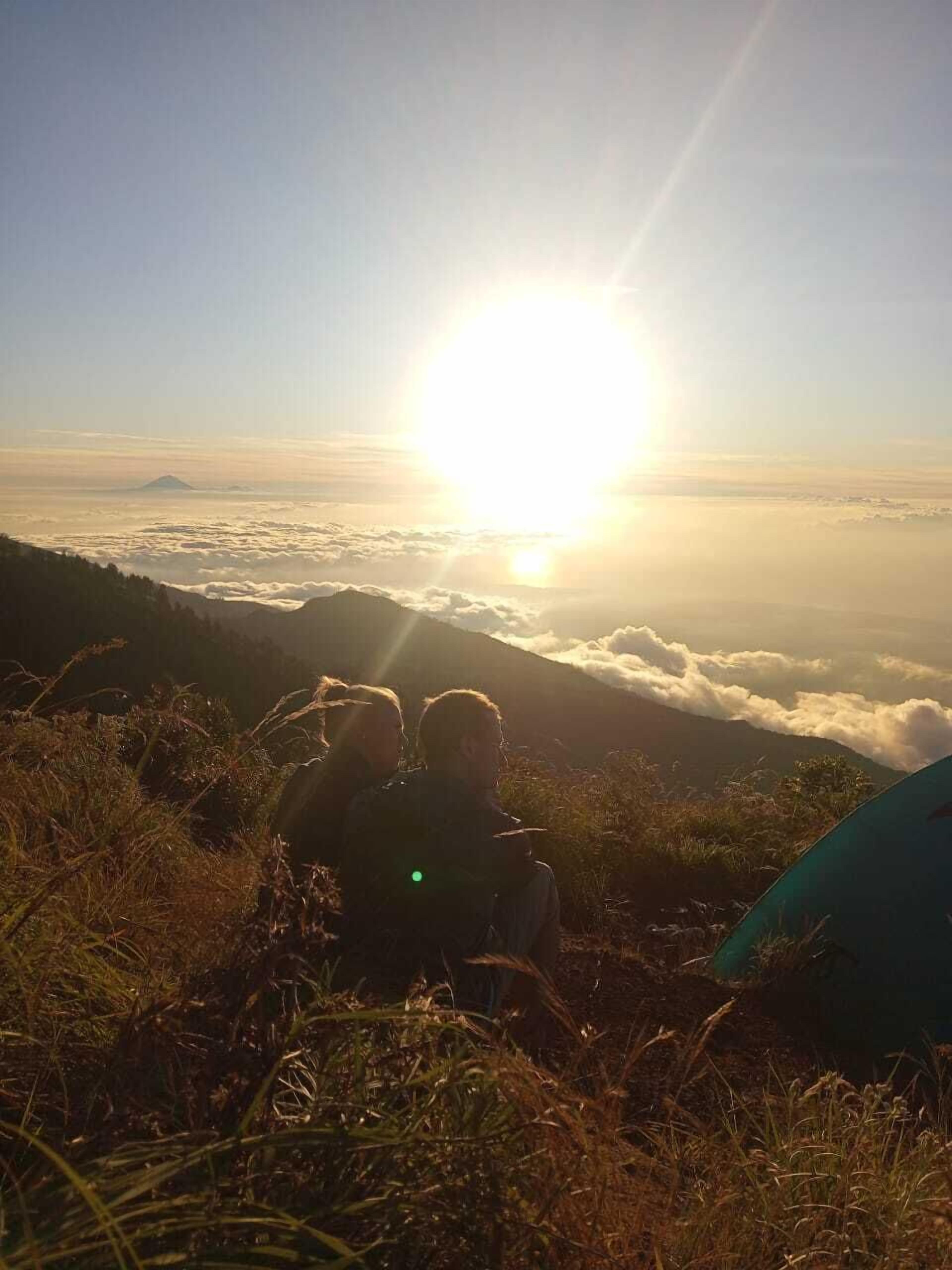 A group of people sitting on a grassy hill overlooking a sea of clouds with the sun shining brightly in the background.