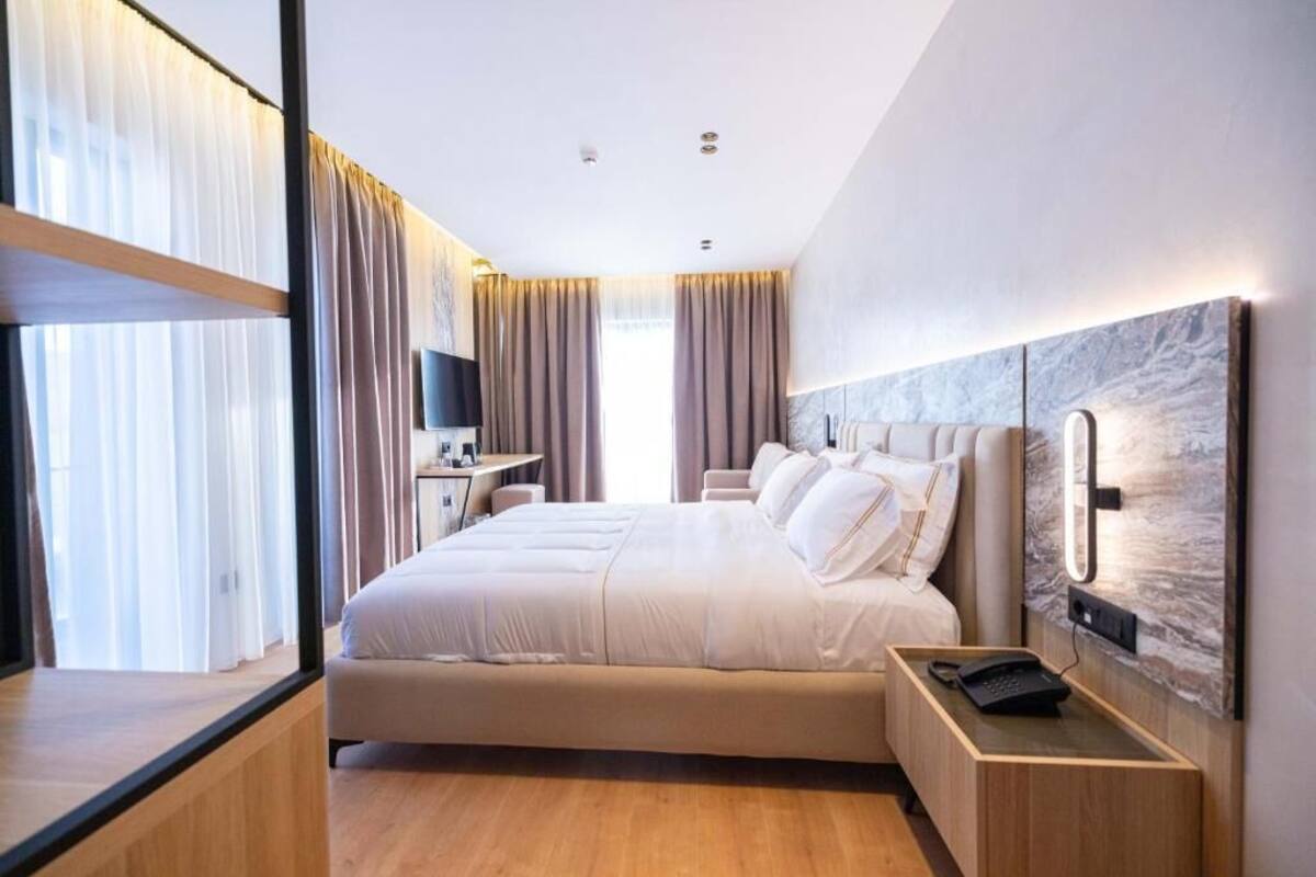 A modern hotel room with a large bed, a bedside table with a phone, a desk with a chair, and a large window with curtains.