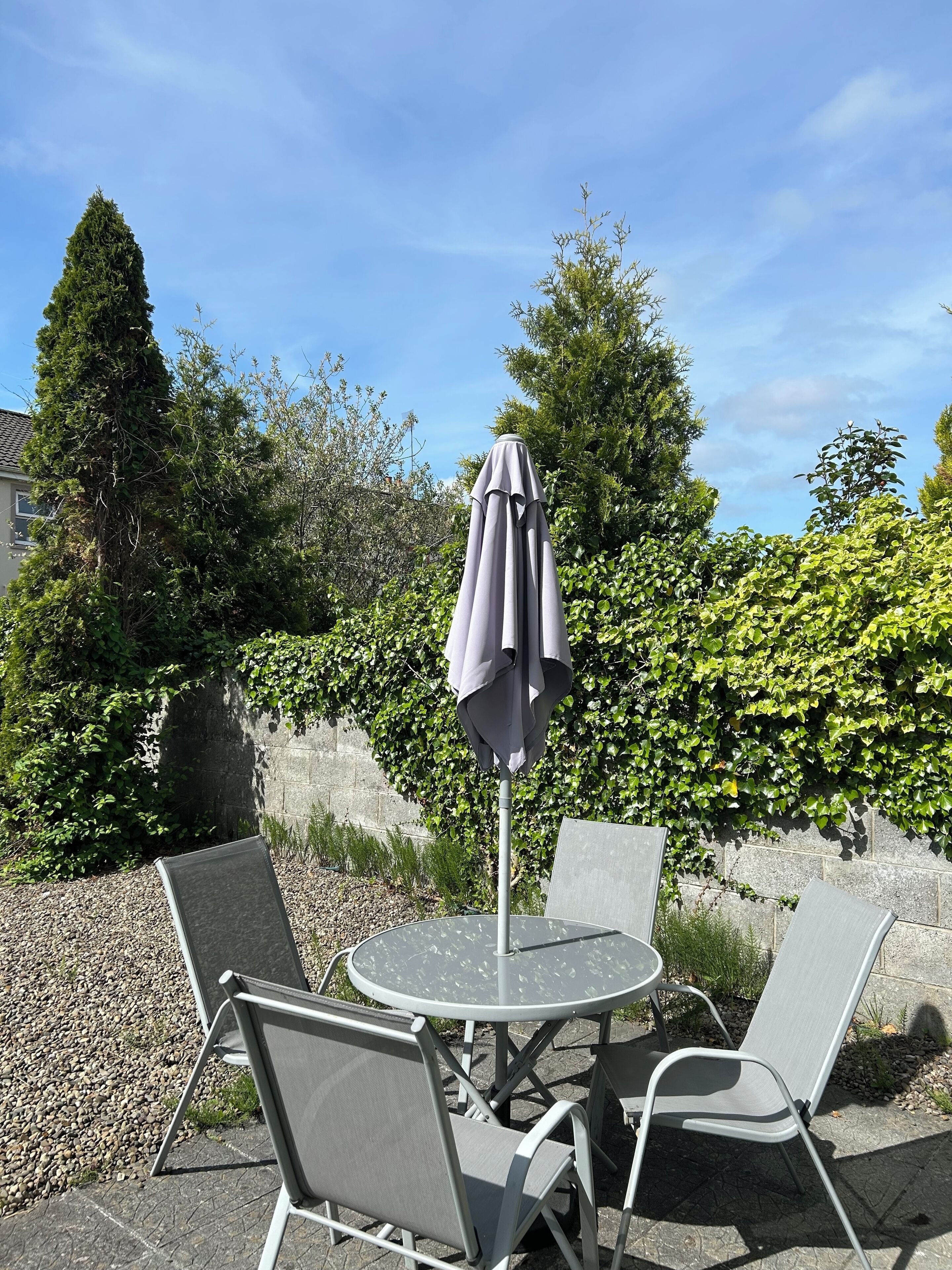 An outdoor seating area with a round table and four chairs, an umbrella, and a hedge in the background.