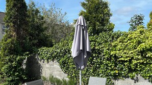 Un espacio al aire libre con una mesa redonda y cuatro sillas, una sombrilla y un seto al fondo.