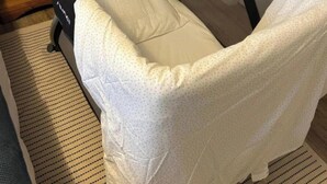 Una cuna para bebé con colchón blanco y estructura negra, ubicada en una habitación con suelo de madera y una puerta blanca al fondo.