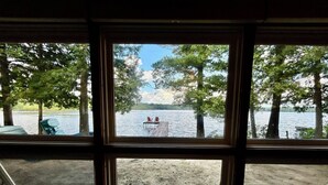 Une vue à travers une fenêtre sur un lac, avec un quai et des chaises, le tout entouré d’arbres.