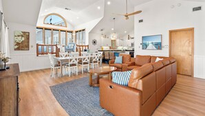 A spacious living room with a large brown leather sofa, a dining area with white chairs, a kitchen in the background, and a view of the outside through large windows. - 6BR Duck Resort | Hot Tub, Beach & Village Walk (Duck)
