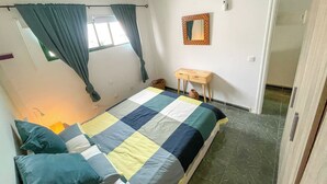 A bedroom with a bed, curtains, a small wooden table, and a mirror on the wall. - Mamarosa Fuerteventura: Family Base 5 min from the Airport + Telework Space (Nuevo Horizonte)