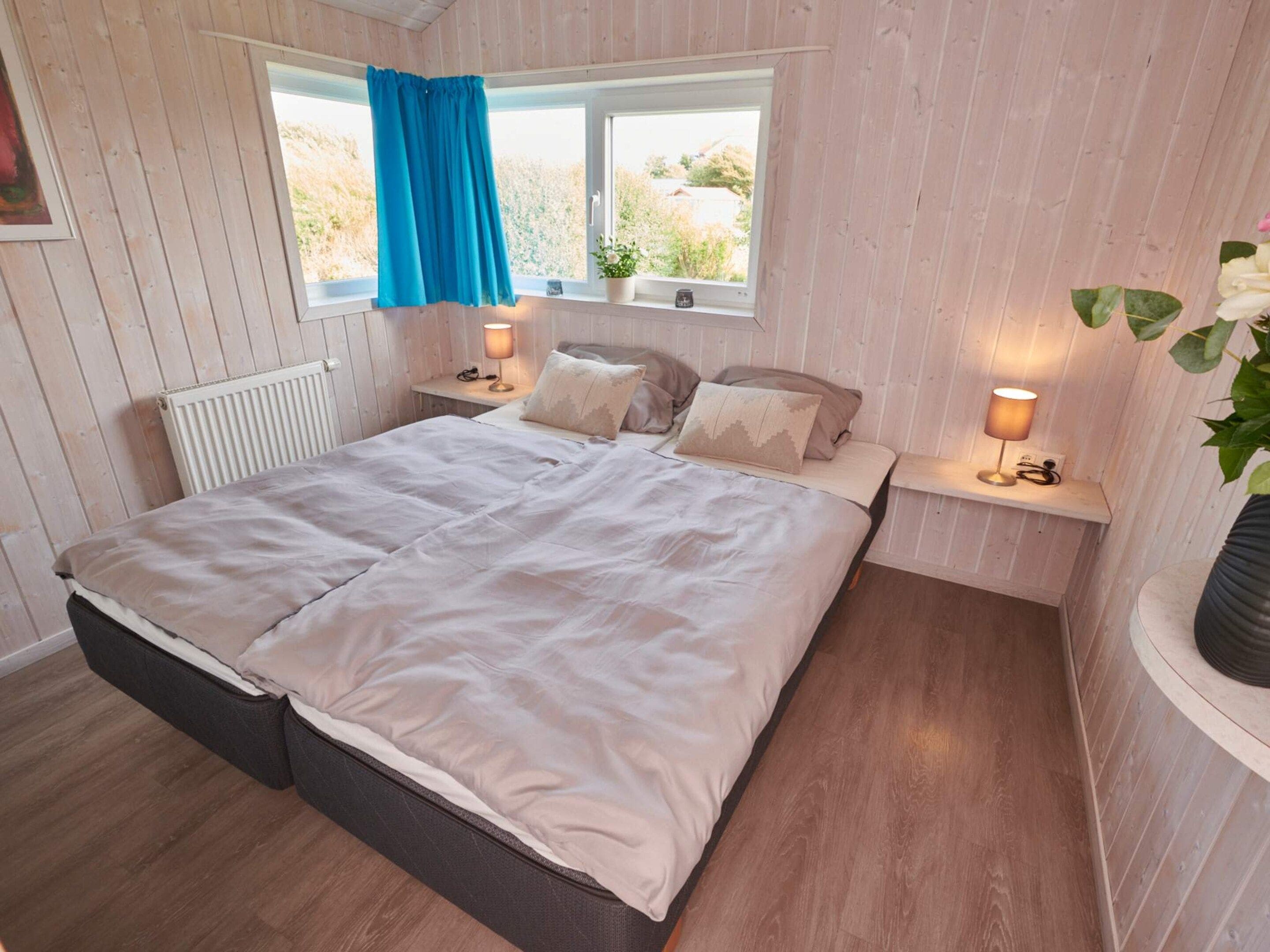 A bedroom with a bed, bedside lamps, a window with curtains, a radiator, and a potted plant.