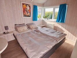 A bedroom with a bed, a bedside table with a lamp, a window with curtains, and a painting on the wall.