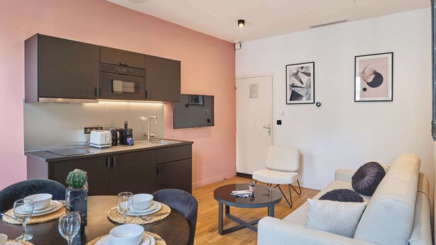 Eine moderne Küche mit dunklen Schränken, einem eingebauten Ofen und einer Kaffeemaschine. Ein Esstisch ist gedeckt mit Tellern, Gläsern und einem Kaktus. Im Wohnbereich befindet sich eine weiße Couch und ein kleiner runder Tisch.