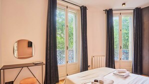Ein Hotelzimmer mit einem Bett, einem runden Spiegel, einem kleinen Tisch und einem Fenster mit Vorhängen.