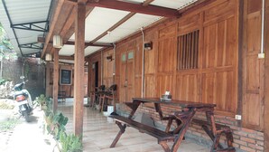 A covered outdoor seating area with wooden tables and chairs, a motorcycle parked outside, and a sign indicating a free stay. - Banyu Kasongan Homestay (Bangunjiwo)