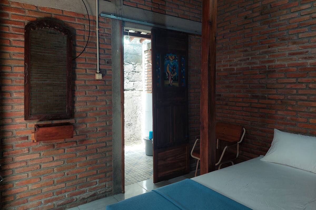 Una habitación con pared de ladrillo, puerta de madera, cama con ropa de cama blanca y un tapete azul.
