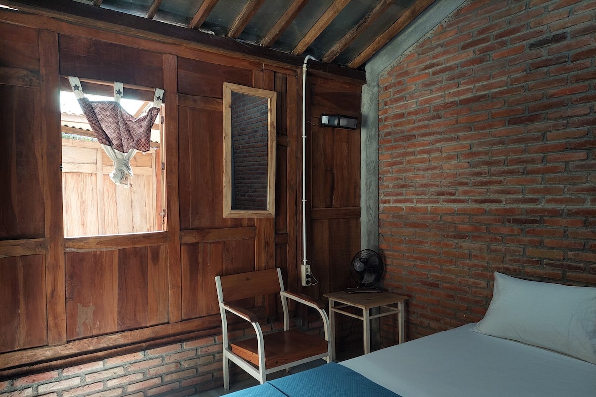 Una habitación con cama, una silla, una mesita, un ventilador y una ventana con cortinas.