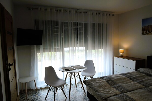 Een hotelkamer met een bed, een bureau met lamp, een aan de muur bevestigde televisie, en een raam met dunne gordijnen.