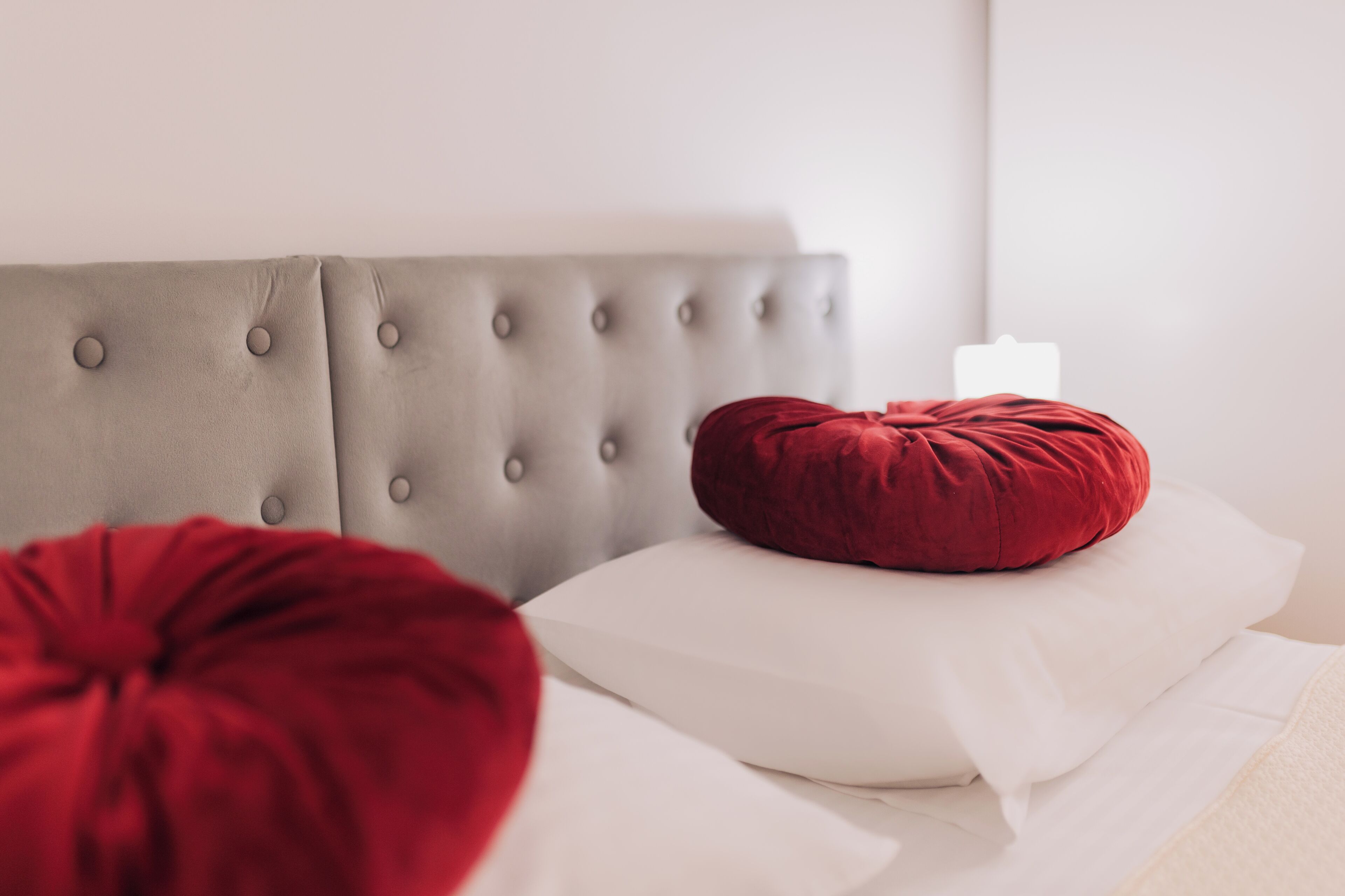 Een bed met een grijs, gestoffeerd hoofdbord, een rood kussen en witte beddengoed.