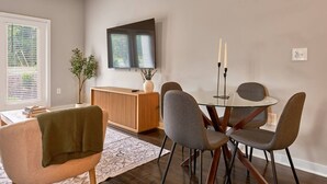Ein Essbereich mit einem runden Glastisch, vier Stühlen, einer Holzkommode, einem Flachbildfernseher und einem Blumentopf.