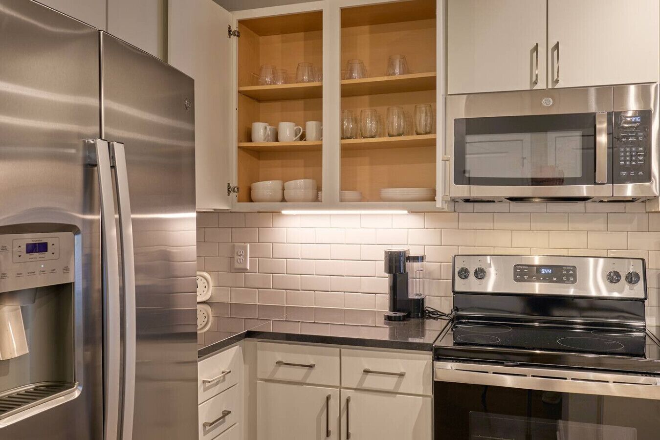 A modern kitchen with stainless steel appliances, including a refrigerator, microwave, and oven. There are wooden cabinets with glassware and dishes.