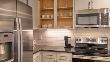 A modern kitchen with stainless steel appliances, including a refrigerator, microwave, and oven. There are wooden cabinets with glassware and dishes.
