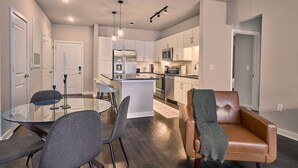 A modern kitchen with white cabinets, stainless steel appliances, and a central island. There is a dining area with a glass table and chairs, and a leather sofa in the living room.