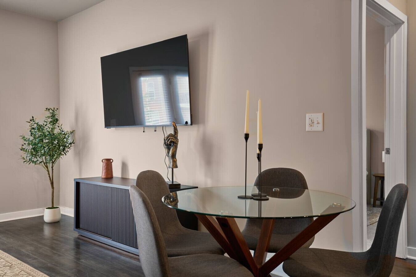 Un comedor moderno con una mesa redonda de vidrio, cuatro sillas, una planta en maceta, una escultura y un televisor de pantalla plana montado en la pared.