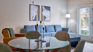 A modern living room with a glass table, blue sofa, and two abstract paintings.