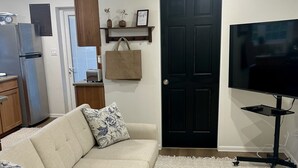 A living room with a beige sectional sofa, a matching ottoman, a black TV stand, and a kitchen area with a refrigerator and a wooden cabinet. - Charming 1-bedroom apartment centrally located to all that Bermuda has to offer (Devonshire)