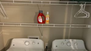 A laundry room with two white front-loading washing machines and a shelf above containing cleaning supplies.