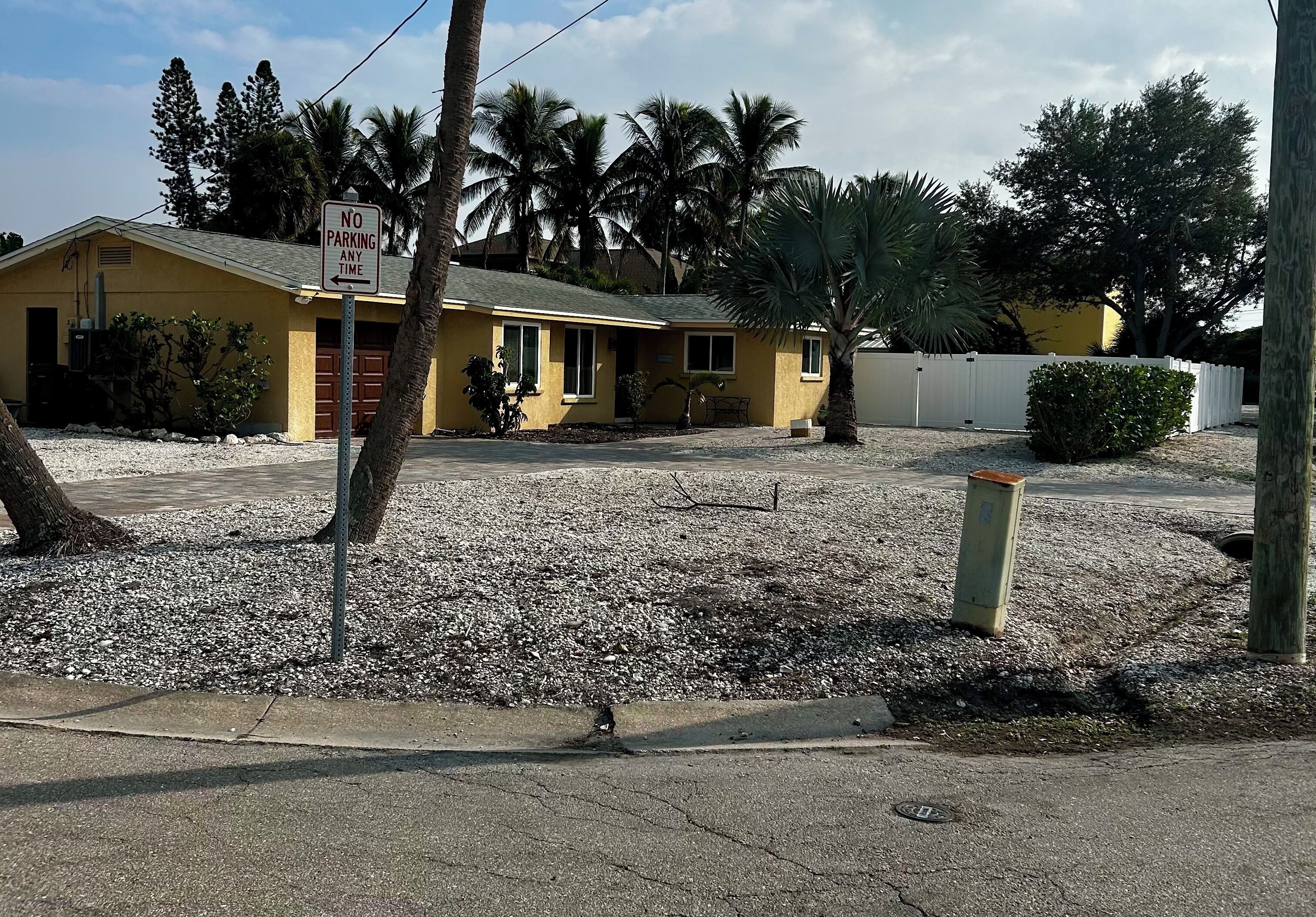 A yellow building with a 'No Parking Any Time' sign, surrounded by palm trees and a gravel driveway.