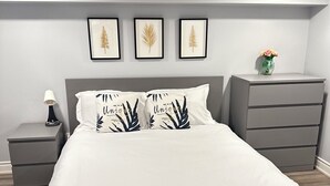 Une chambre à coucher avec un lit, des tables de chevet grises, une commode et des reproductions botaniques encadrées accrochées au mur.