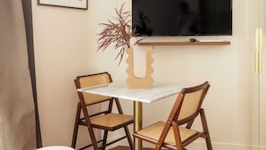 A small dining area with a white table, two wooden chairs, a round pink stool, a TV mounted on the wall, and a framed artwork. - Port-royal Residence-magnificent Apartment -1br-4p (Paris)